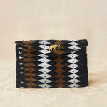 A small beaded rectangular clutch bag with a black, brown, and white geometric diamond pattern and a small gold elephant charm on the front flap, set against a neutral beige background.