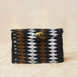 A small beaded rectangular clutch bag with a black, brown, and white geometric diamond pattern and a small gold elephant charm on the front flap, set against a neutral beige background.