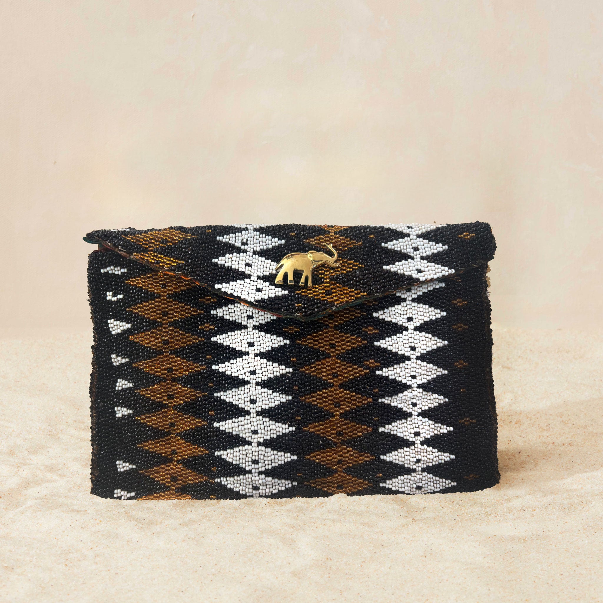 A small beaded rectangular clutch bag with a black, brown, and white geometric diamond pattern and a small gold elephant charm on the front flap, set against a neutral beige background.