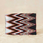 Back view of a beaded clutch purse featuring a bold chevron pattern in black, white, red, and brown, with a small gold elephant charm on the front flap, on a neutral beige background.
