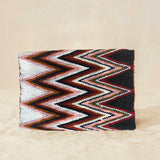 Back view of a beaded clutch purse featuring a bold chevron pattern in black, white, red, and brown, with a small gold elephant charm on the front flap, on a neutral beige background.