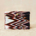 A beaded clutch purse featuring a bold chevron pattern in black, white, red, and brown, with a small gold elephant charm on the front flap, on a neutral beige background.