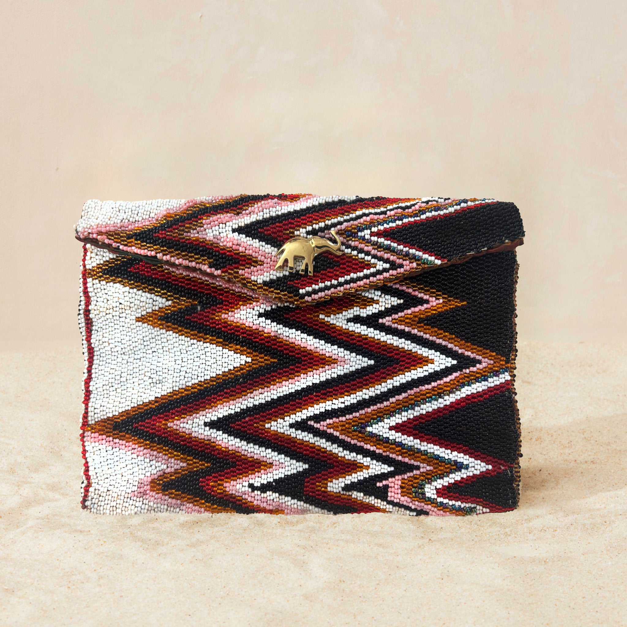 A beaded clutch purse featuring a bold chevron pattern in black, white, red, and brown, with a small gold elephant charm on the front flap, on a neutral beige background.