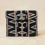 A small beaded clutch bag with a geometric pattern in black, brown, and white, featuring a small gold elephant clasp on the front flap, set against a neutral beige background.