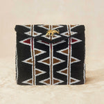A black beaded clutch purse featuring a geometric pattern of white, bronze and red triangles, with a small gold elephant charm on the front flap, set against a neutral beige background.