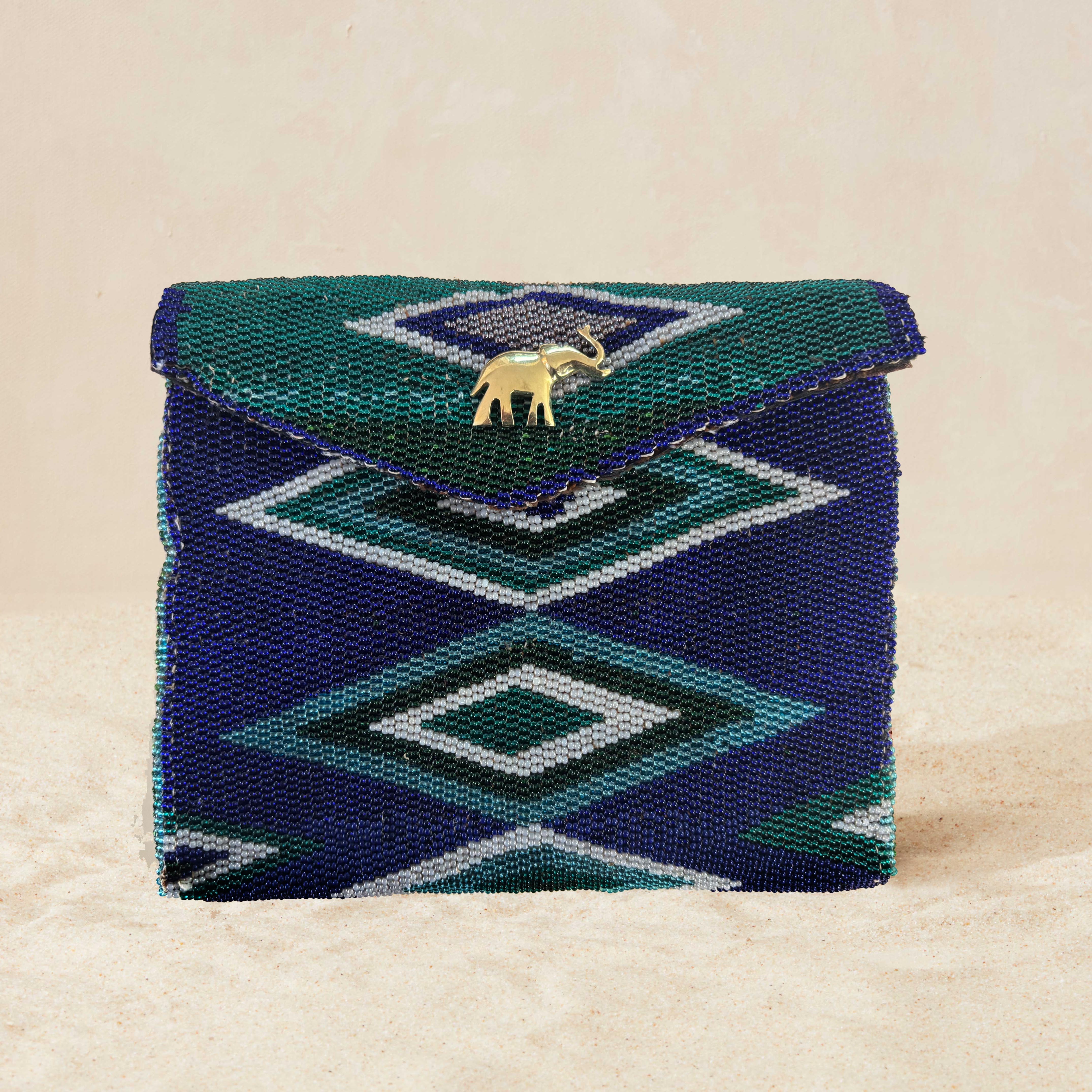 Blue, green, teal, turquoise geometric beaded clutch bag with a brass elephant charm on a beige sand background