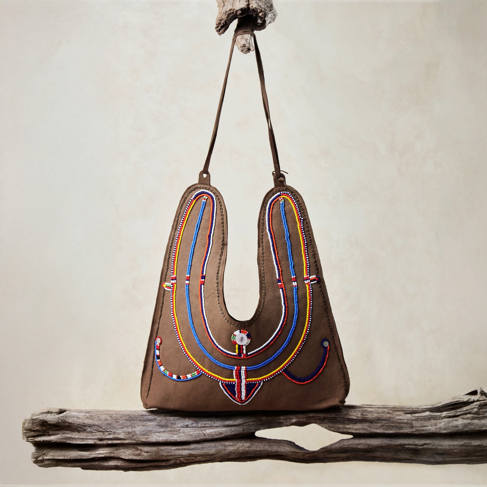 Caramel brown leather suede handbag with colorful hand beading hanging on a wooden branch against a beige background