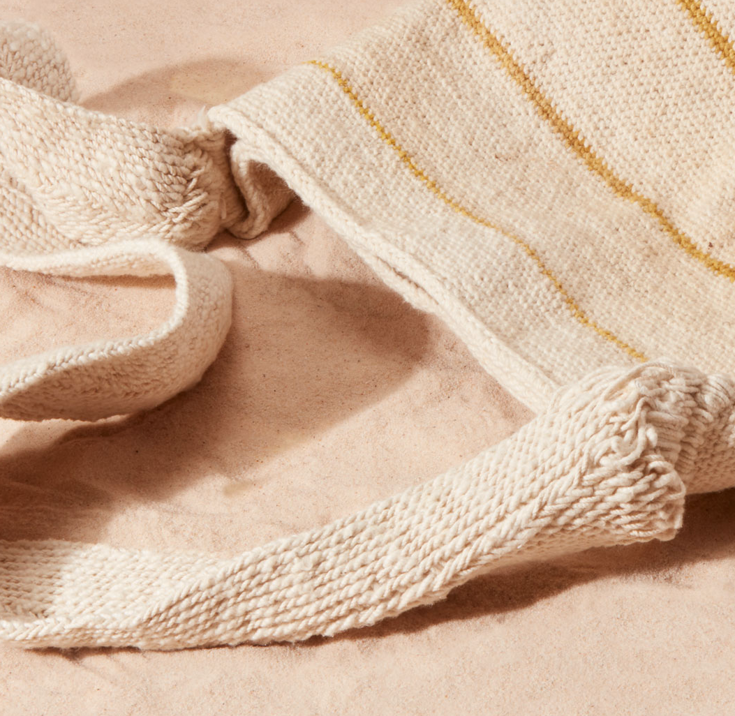Detail view of woven straps of woven ivory and yellow striped bag against a sand background