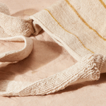 Detail view of woven straps of woven ivory and yellow striped bag against a sand background