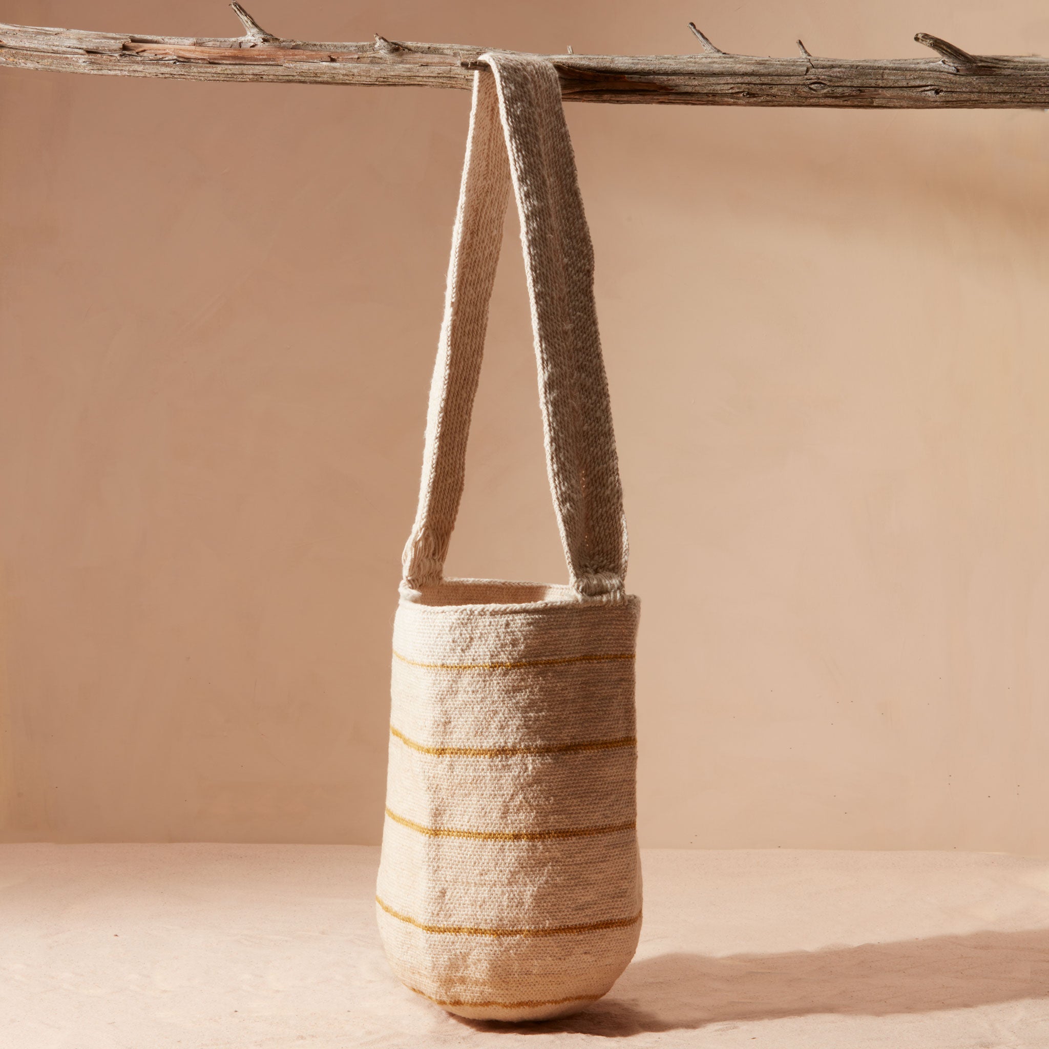 Woven bag with a strap and thin yellow/brown stripes hanging on a branch against a beige background