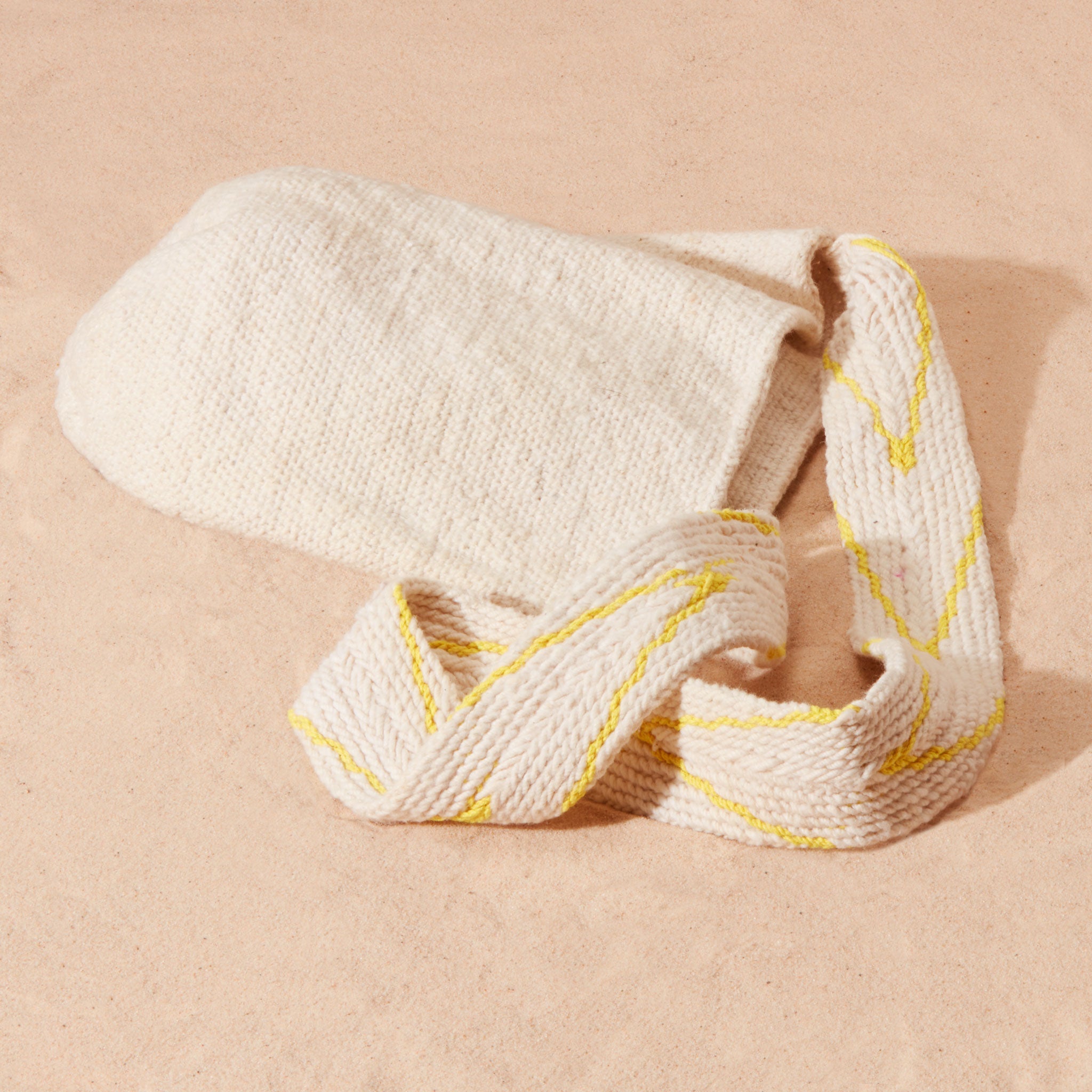 Flat view of ivory woven bag with yellow and beige v patterned strap on a sand surface
