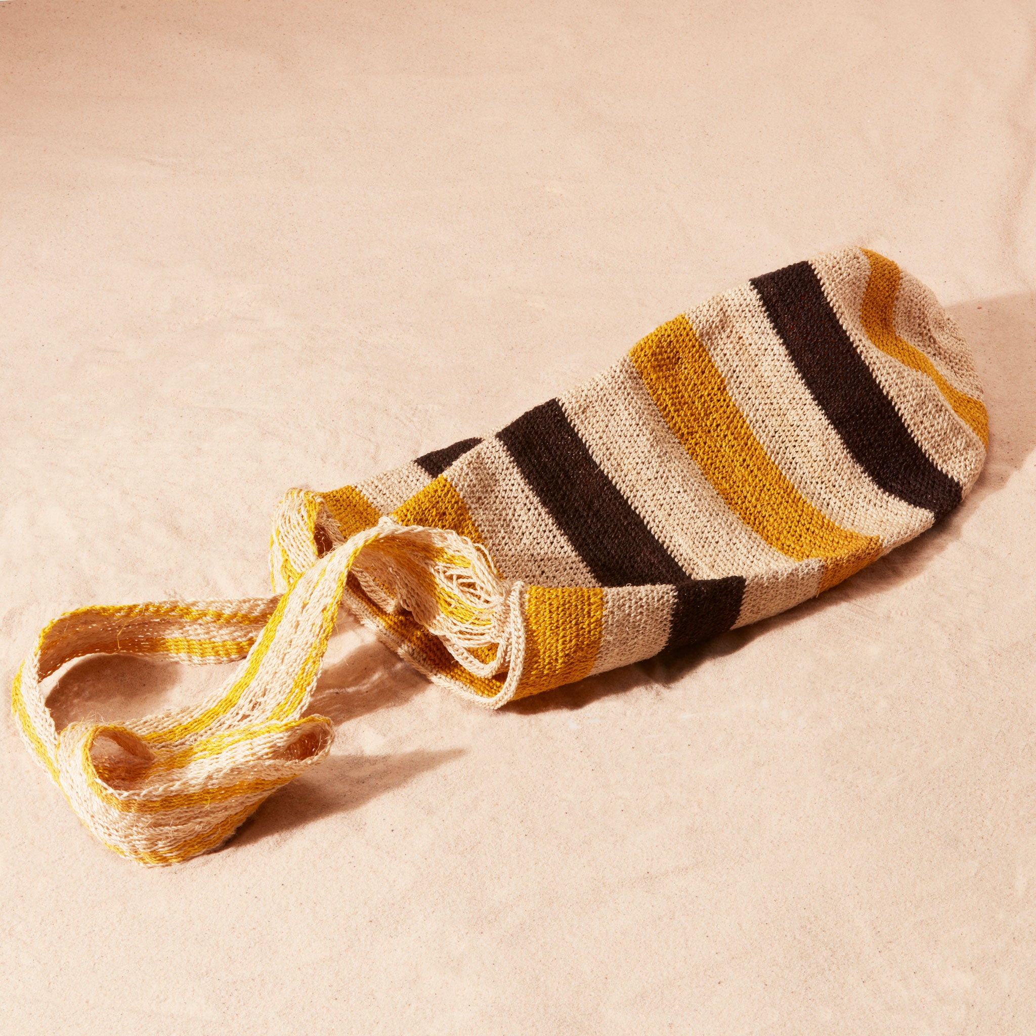 Flat view of black, yellow and ivory thick striped woven bag with a loose weave strap in ivory and yellow on a sand background