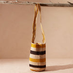 Yellow, black and ivory striped woven  handbag with a strap hanging on a branch on a beige background