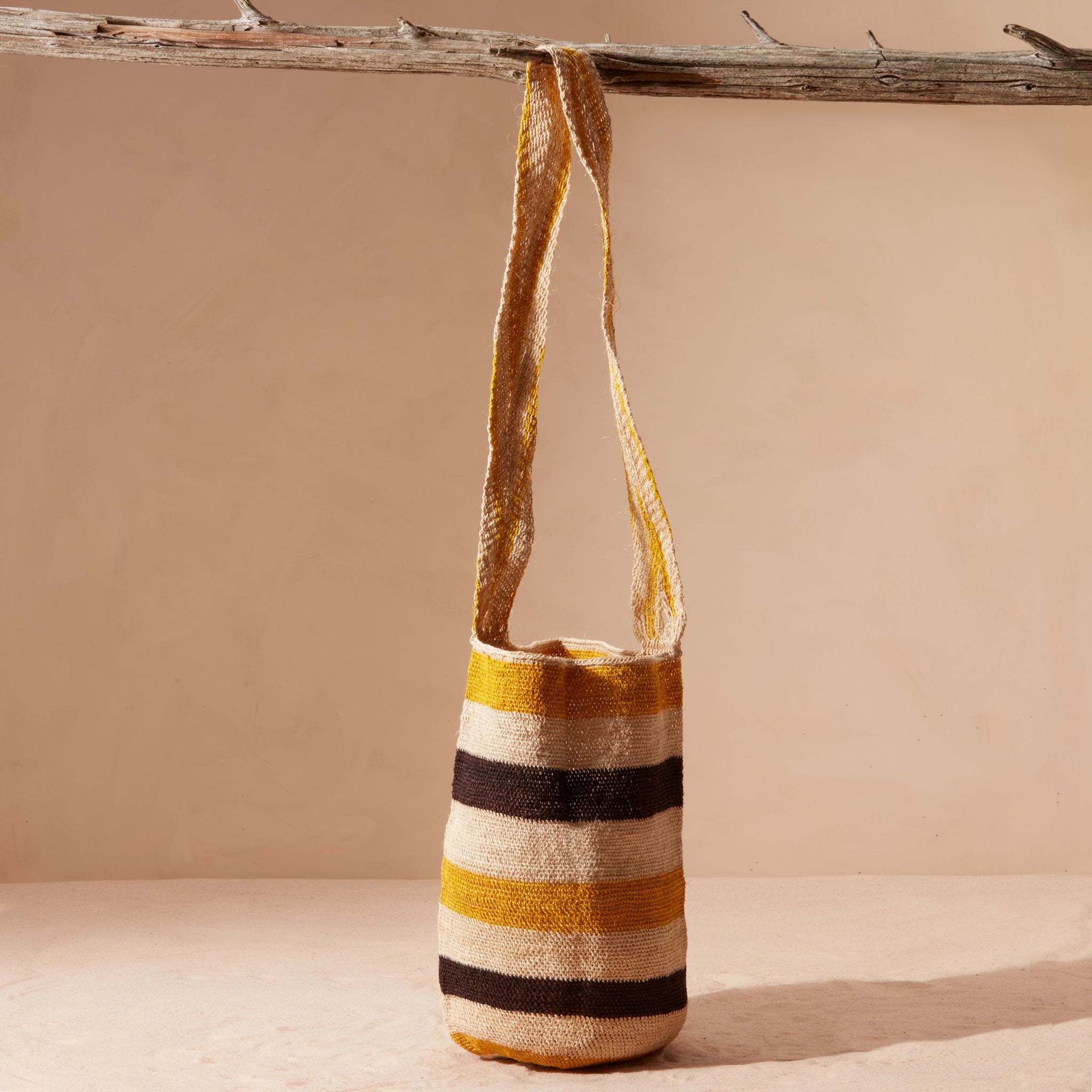 Yellow, black and ivory striped woven  handbag with a strap hanging on a branch on a beige background