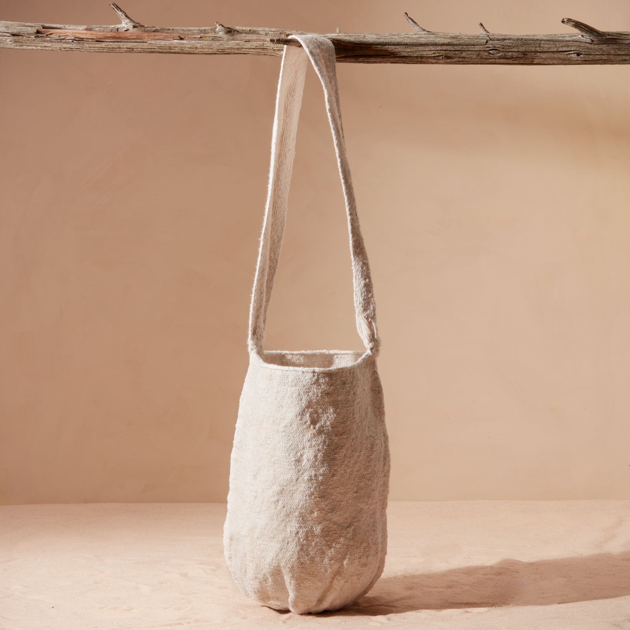 Textured ivory woven bag hanging from the strap from a branch against a beige background