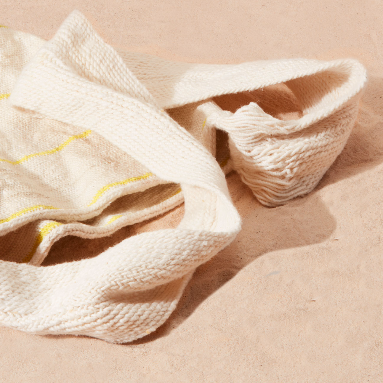 Detail view of woven ivory strap of the woven bag with thin yellow stripes against a sand background