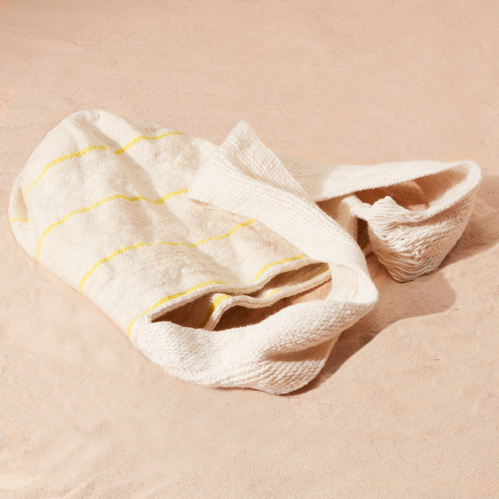 Flat view of ivory woven bag with thin yellow stripes on a sand background