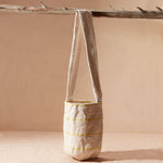 Woven ivory bag with thin yellow stripes with strap hanging from a branch on a beige background