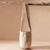Woven ivory bag with thin yellow stripes with strap hanging from a branch on a beige background