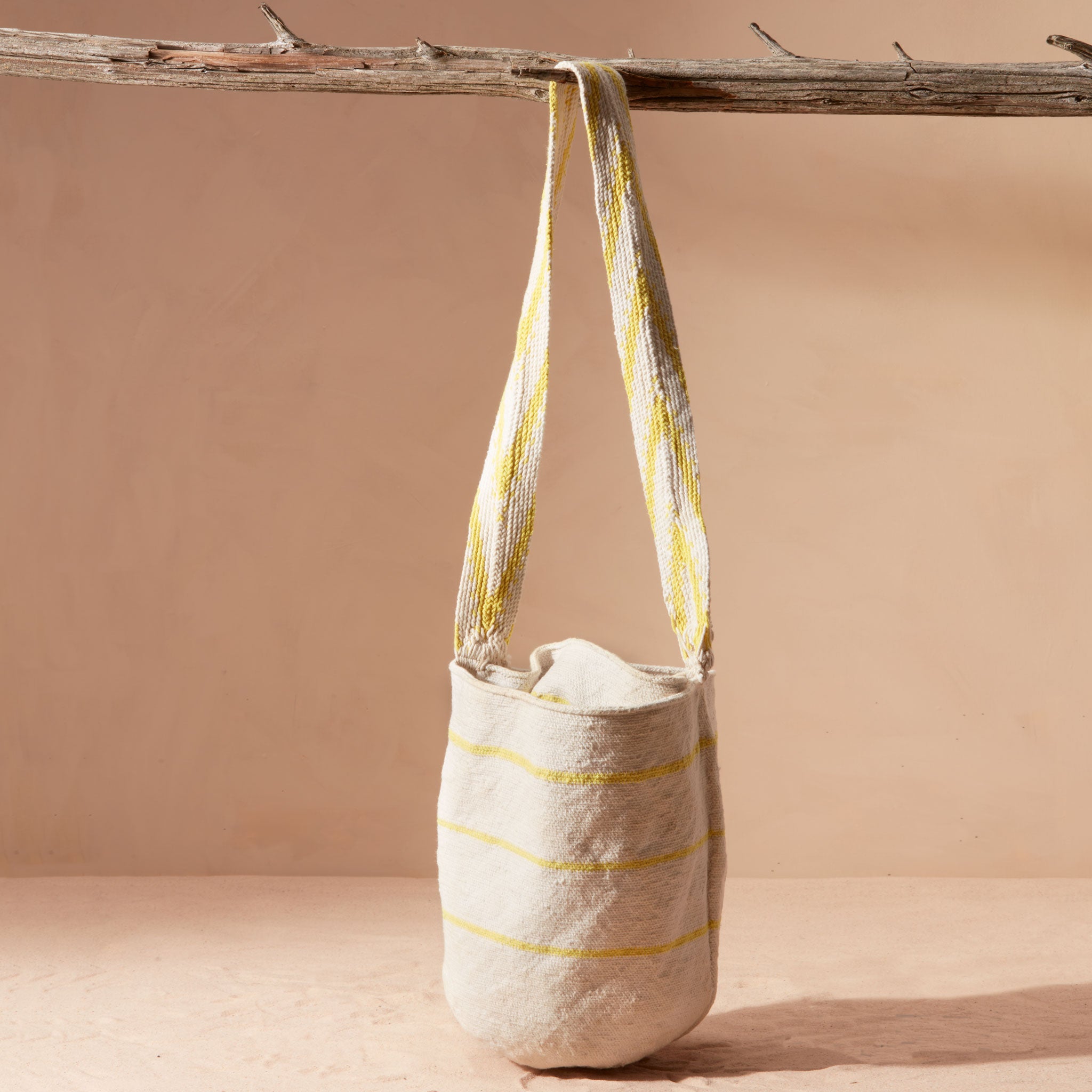 Ivory woven bag with yellow stripes hanging from a branch against a beige background