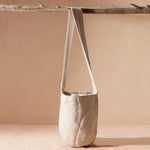 Ivory woven bag with strap hanging from a branch against a beige background
