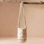 Woven ivory bag with thin yellow/brown and black stripes hanging from the strap from a branch against a beige background