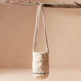 Woven ivory bag with thin yellow/brown and black stripes hanging from the strap from a branch against a beige background