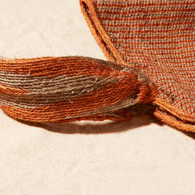 Close up view of Brown-orange and grey woven bag with strap on a sand background