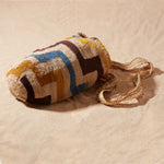 Side view of hand-woven bag with brown, yellow, blue, ivory geometric pattern on a sand background