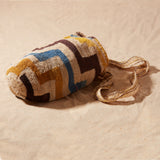 Side view of hand-woven bag with brown, yellow, blue, ivory geometric pattern on a sand background