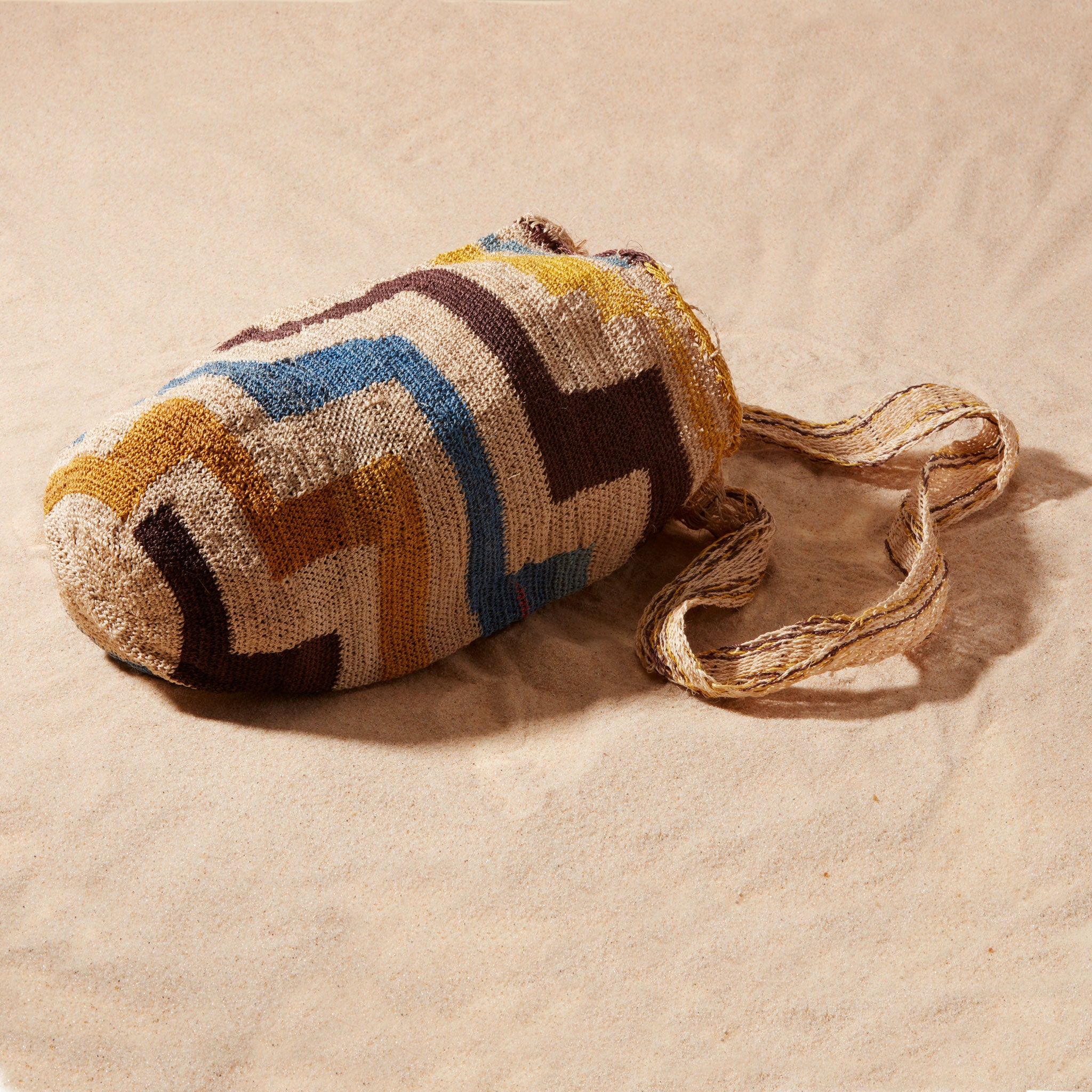 Side view of hand-woven bag with brown, yellow, blue, ivory geometric pattern on a sand background