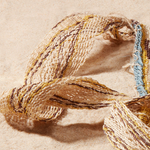 Close up view of very loosely woven strap of woven bag with brown, yellow, blue, ivory geometric pattern on a sand background