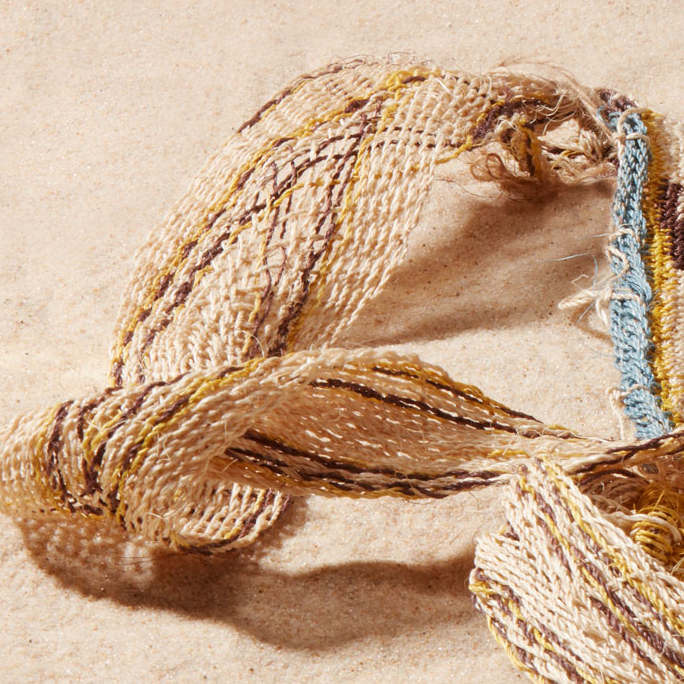 Close up view of very loosely woven strap of woven bag with brown, yellow, blue, ivory geometric pattern on a sand background
