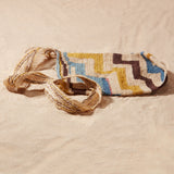Flat view of hand-woven bag with brown, yellow, blue, ivory geometric pattern against a sand background