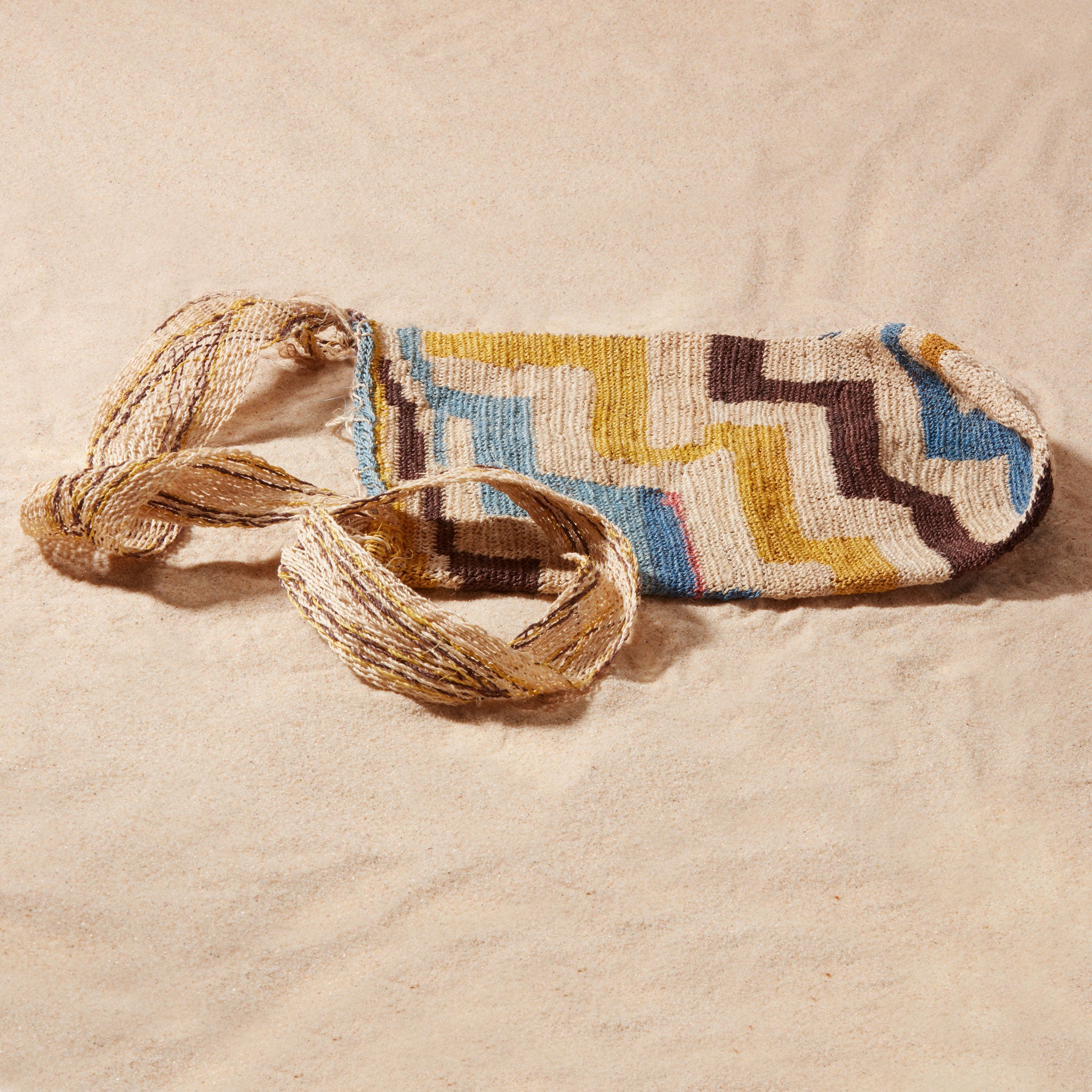 Flat view of hand-woven bag with brown, yellow, blue, ivory geometric pattern against a sand background