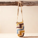 Hand-woven bag with brown, yellow, blue, ivory geometric pattern hanging on a wooden branch against a beige background