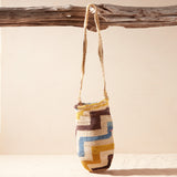 Hand-woven bag with brown, yellow, blue, ivory geometric pattern hanging on a wooden branch against a beige background