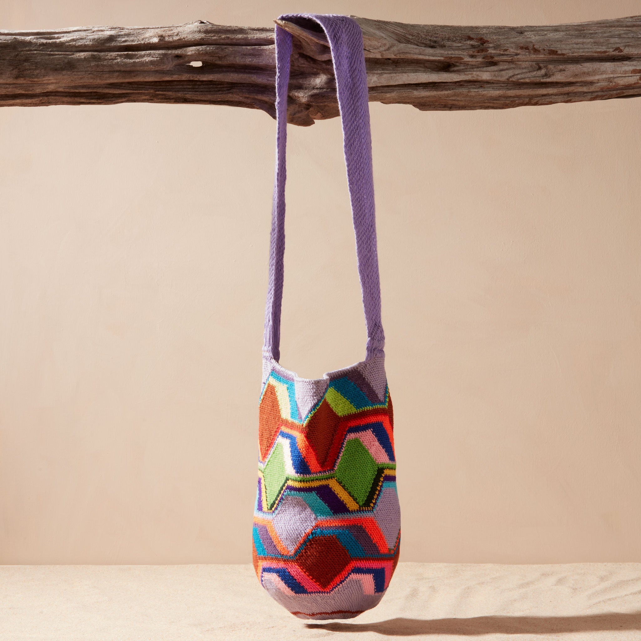 Colorful geometric-patterned bag with a purple strap hanging on a wooden branch against a beige background