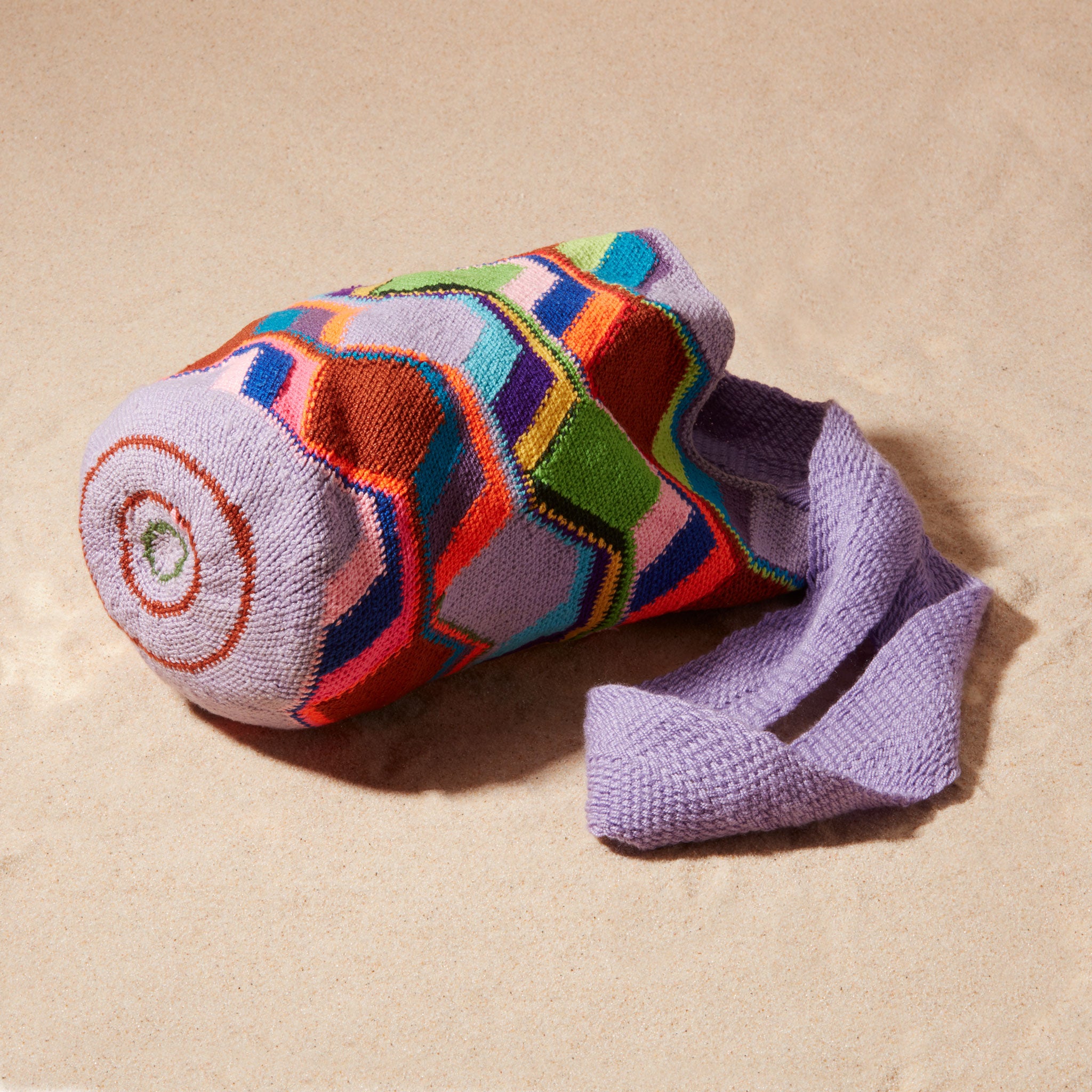 Bottom view of colorful geometric-patterned bag with a purple strap on a sand surface