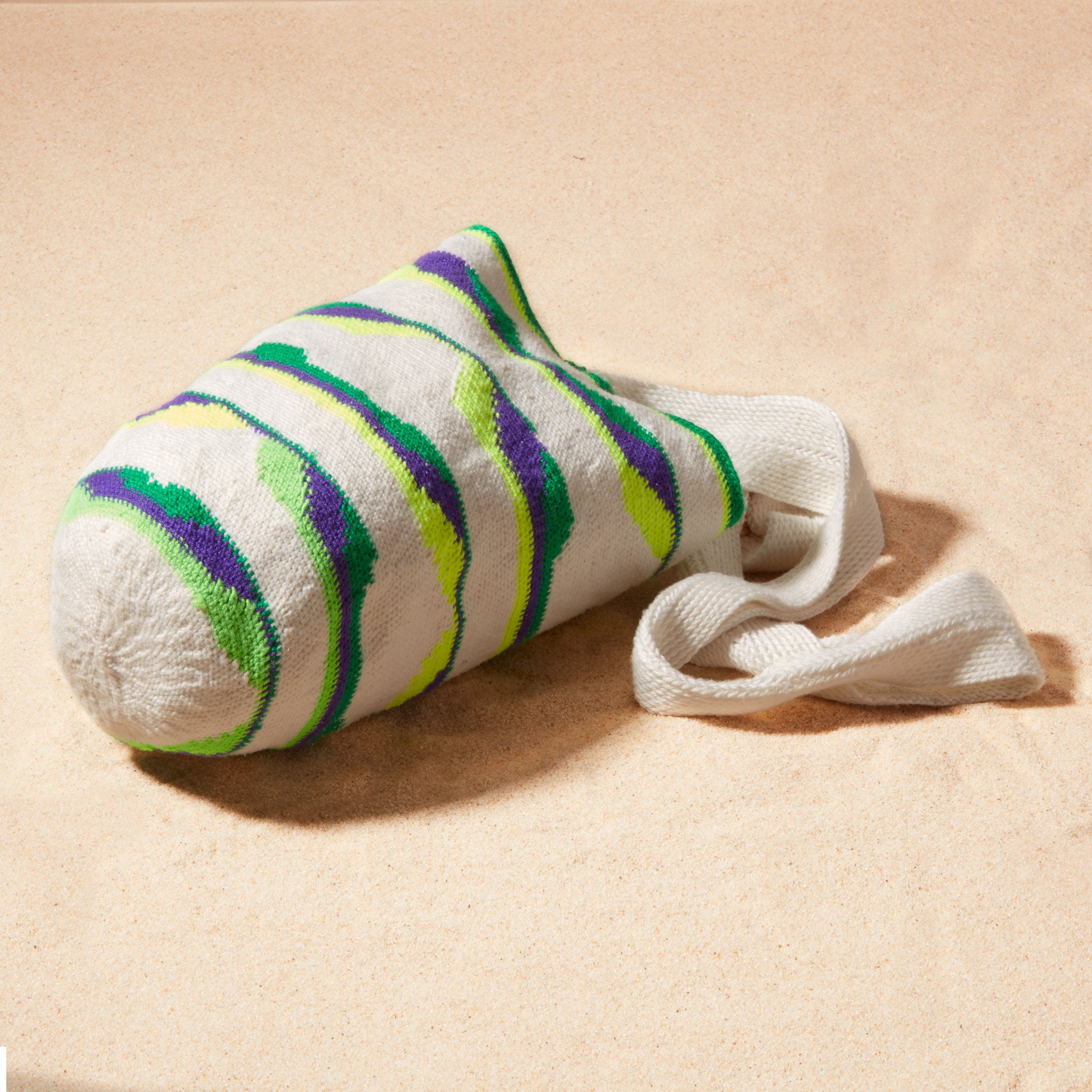 Side view of ivory, green, neon green, purple woven bag on a sand background