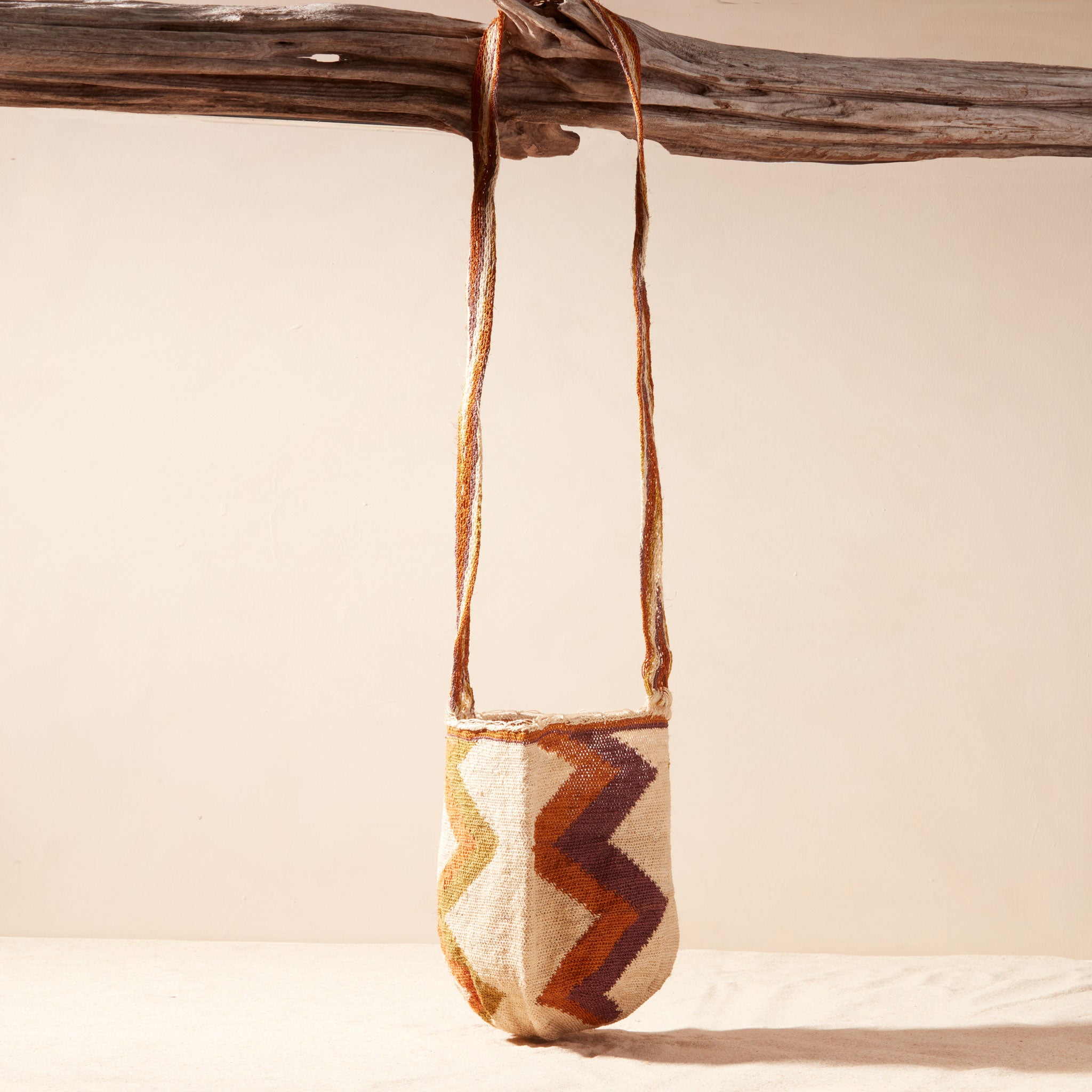 Textured woven bag with a zigzag pattern hanging from a branch against a beige background