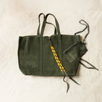 Flat view of olive green suede leather tote bag with yellow, black, and red beaded tassel ties including an attached interior zipper pouch on a sand background