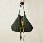 Olive green suede leather tote bag with yellow, black, and red beaded tassel ties hanging on a branch with a beige background