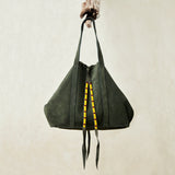 Olive green suede leather tote bag with yellow, black, and red beaded tassel ties hanging on a branch with a beige background