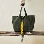 Olive green suede leather tote bag with yellow, black, and red beaded tassel ties hanging and sitting on wooden branches with a sand background