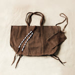 Flat view of brown sand suede leather tote bag with white, black, and red beaded tassel ties including an attached interior zipper pouch on a sand background