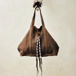 Brown sand suede leather tote bag with white, black, and red beaded tassel ties handing on a branch with a beige background