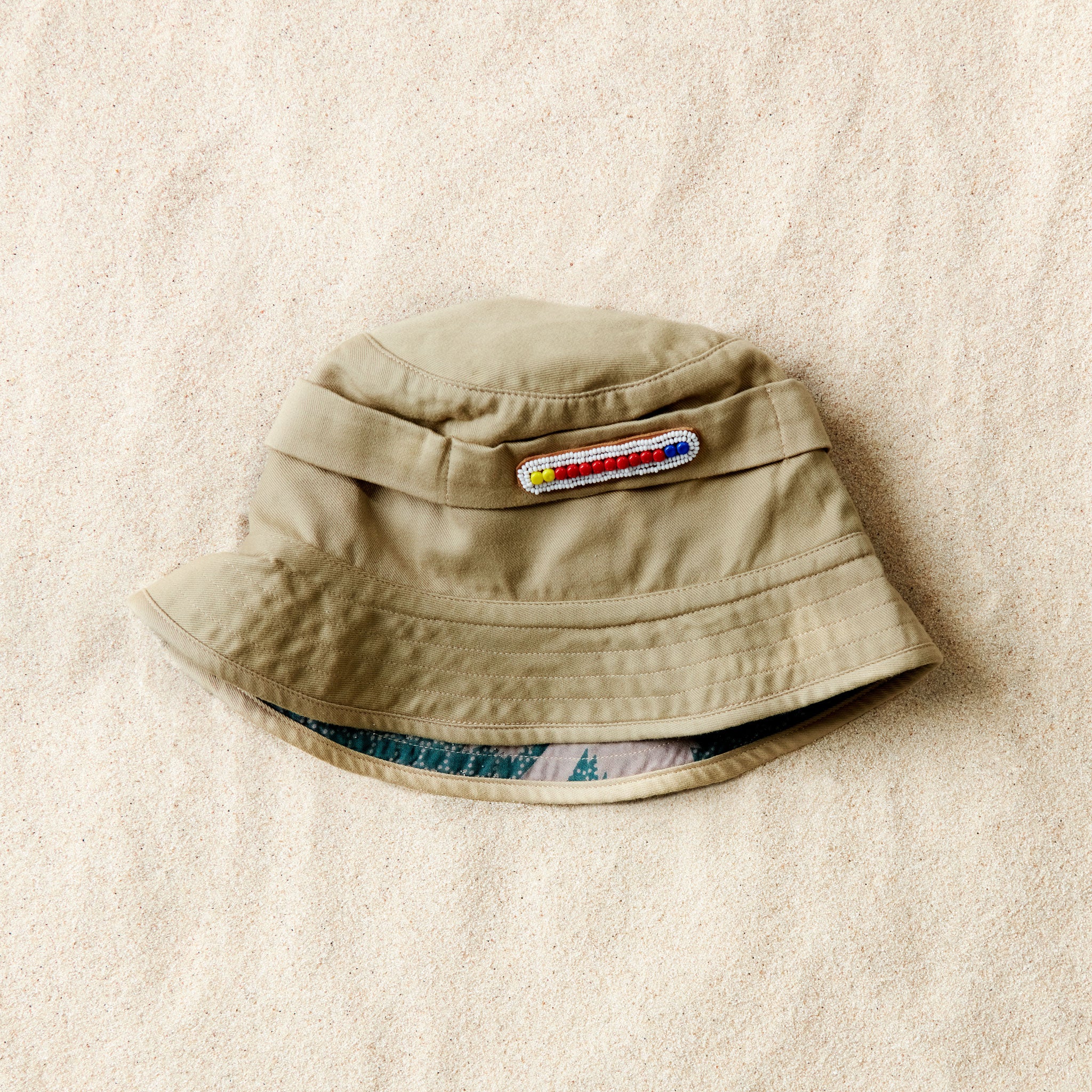 Flat view of khaki bucket hat with a white and colorful beaded patch and colorful kanga fabric lining on a sand background