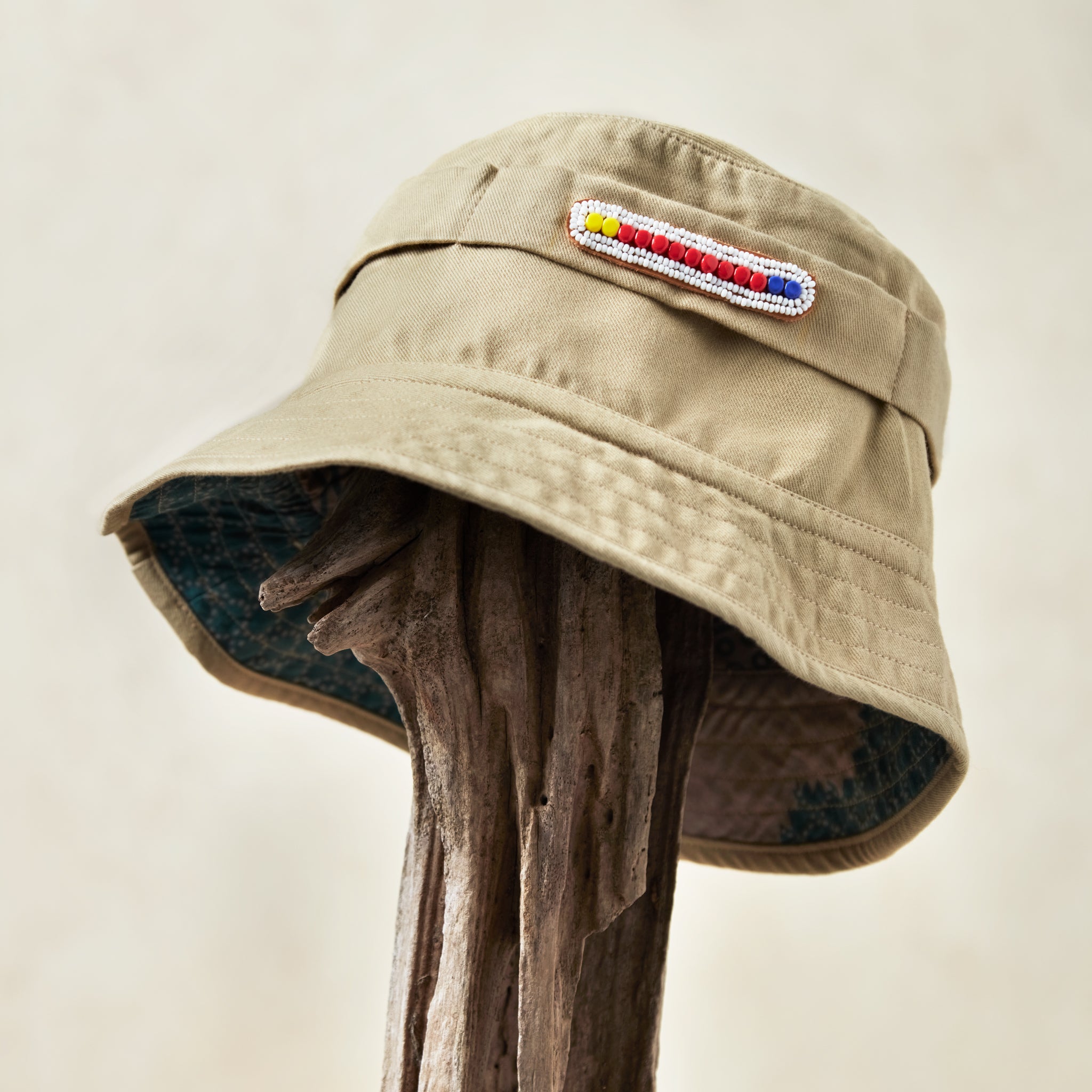 Khaki bucket hat with a white and colorful beaded patch and colorful kanga fabric lining on a wooden post against a beige background