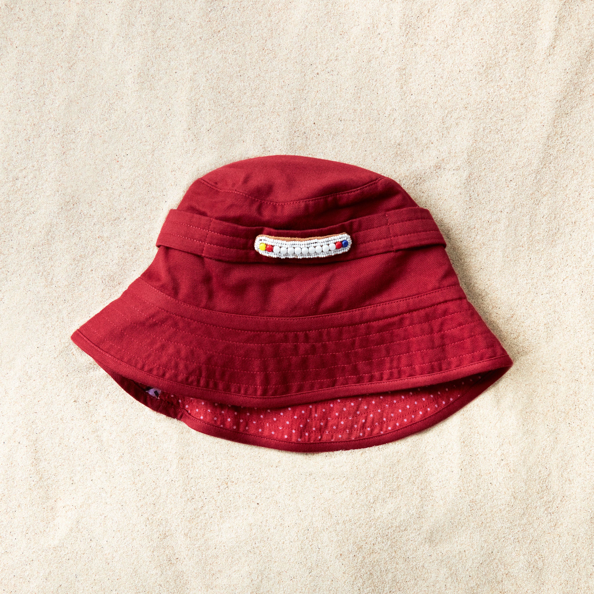 Flat view of red bucket hat with a white and colorful beaded patch on a sand background